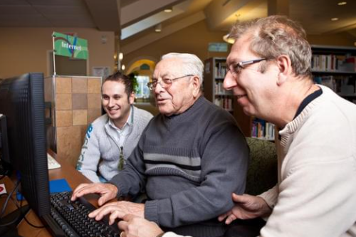 older gentlemen uses a desktop computer while two men look on