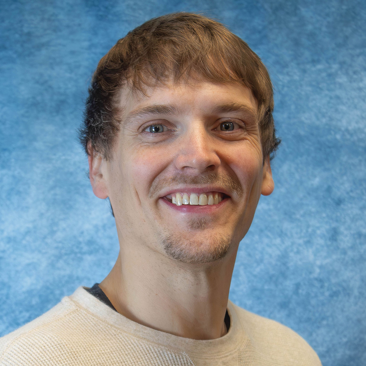 Travis Johnson headshot. Whiteman with sandy brown hair and with a tan long sleave in front of a blue backdrop.